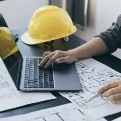 Inżynier analizuje plany, porównując powierzchnię zabudowy z użytkową. Na biurku laptop, żółty kask i rysunki techniczne.