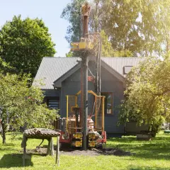 Wiertnica pracuje na działce, budując studnię. W tle widać dom i drzewa.