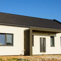 Nowy dom z ciemnym dachem i białymi ścianami, gotowy do odbioru. Widać drzwi wejściowe i okna.