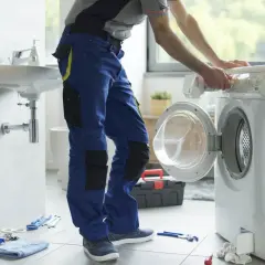 Instalator w niebieskich spodniach pracuje przy pralce, pokazując, jak podłączyć pralkę. Narzędzia leżą na podłodze.