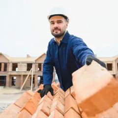 Murarz w białym kasku i niebieskim kombinezonie podnosi cegłę na budowie. W tle widać domy w budowie.