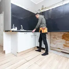 Mężczyzna w pasie narzędziowym planuje remont kuchni od czego zacząć, stojąc przy częściowo zdemontowanej zabudowie.
