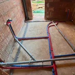 Rozpoczynająca się instalacja wodna w domu, z czerwonymi i niebieskimi rurami biegnącymi po podłodze i ścianach z cegły.