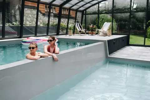 Dwie kobiety relaksują się w basenie infinity pod przeszklonym dachem. W tle widać leżaki i otaczającą zieleń.