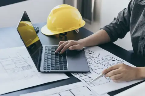 Inżynier analizuje plany, porównując powierzchnię zabudowy z użytkową. Na biurku laptop, żółty kask i rysunki techniczne.