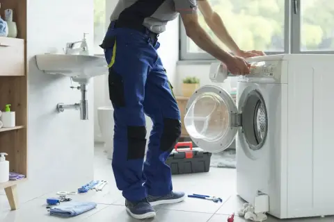 Instalator w niebieskich spodniach pracuje przy pralce, pokazując, jak podłączyć pralkę. Narzędzia leżą na podłodze.