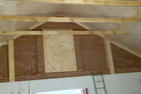 Drewniane belki tworzą konstrukcję dachu wielospadowego nad ceglaną ścianą. Na ścianie widoczny panel z desek ułożonych we wzór jodełki.