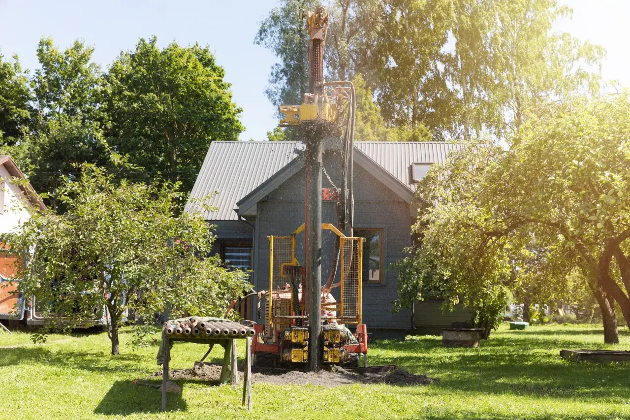 Wiertnica pracuje na działce, budując studnię. W tle widać dom i drzewa.