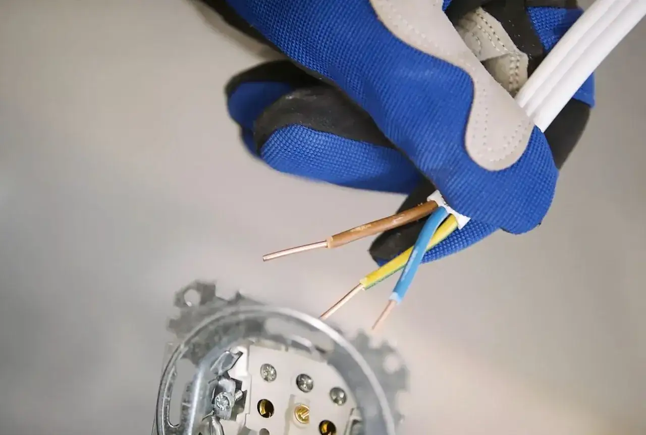 Rękawica pokazuje, jak podłączyć gniazdko elektryczne. Widoczne przewody: brązowy, niebieski i żółto-zielony.