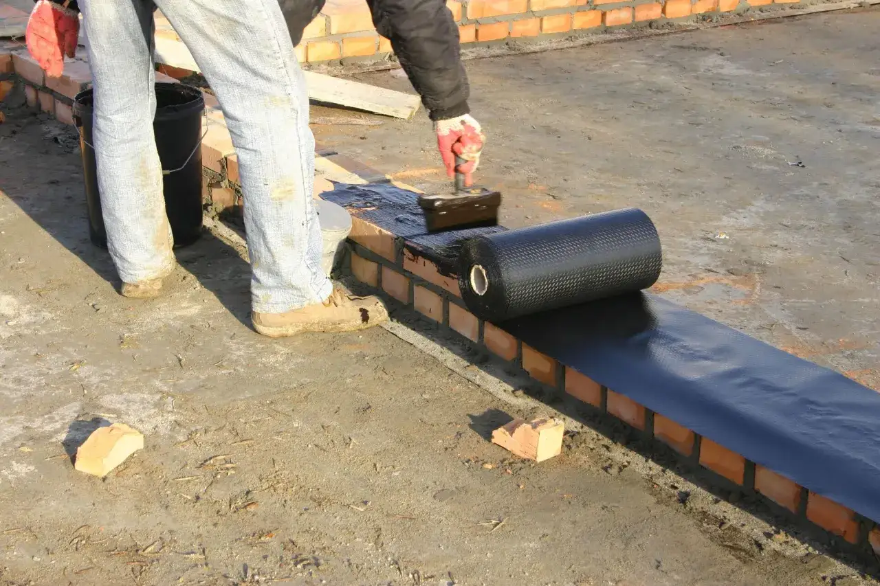 Pracownik z wałkiem rozprowadza hydroizolację na murze. Materiał ma wzór, który zapobiega jego rozwarstwianiu.